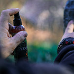 A man's hand spraying his gear with hunting spray. The spray mist is also visible in the image.