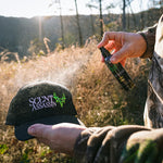 a man's hands with a scent control spray in one hand and a hunting cap in the other. The man is spraying the hunting cap to make it scent-free.